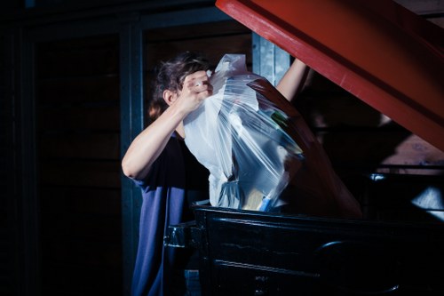A person with short dark hair, wearing a dark blue shirt, is seen disposing of a clear plastic bag filled with various waste materials into a large black rubbish bin. The bin is positioned outdoors on what appears to be a driveway or paved area adjacent to a building with dark wooden cladding and a blue-painted wooden frame. The person's hand is grabbing the top of the plastic bag, which contains mixed refuse such as paper, plastic, and other household waste. The scene suggests a rubbish collection or clearance activity, typical of a professional waste removal service in Brixton, with the background environment appearing shaded and the lighting focused on the individual and their task, supporting an efficient rubbish disposal operation.